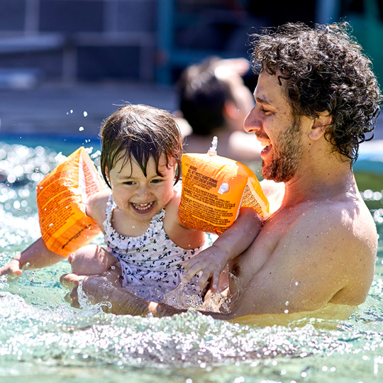 Photo of a father in a pool with a toddler wearing orange swim floaties smiling and splashing the water